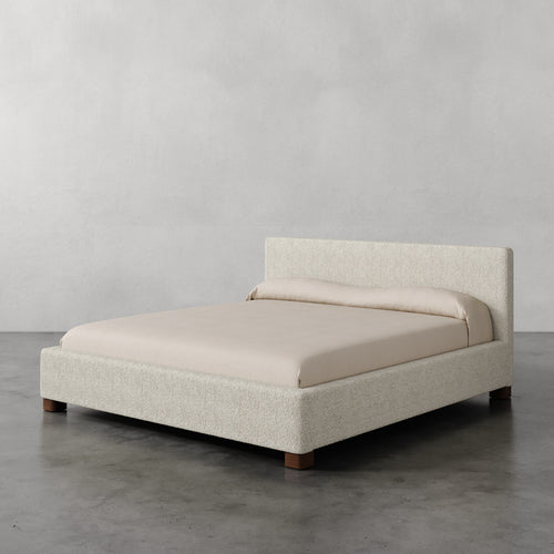 Lemieux et Cie Gérard Bed in ivory bouclé, low-profile platform bed with wrapped headboard and rounded block feet, shown on polished concrete floor.