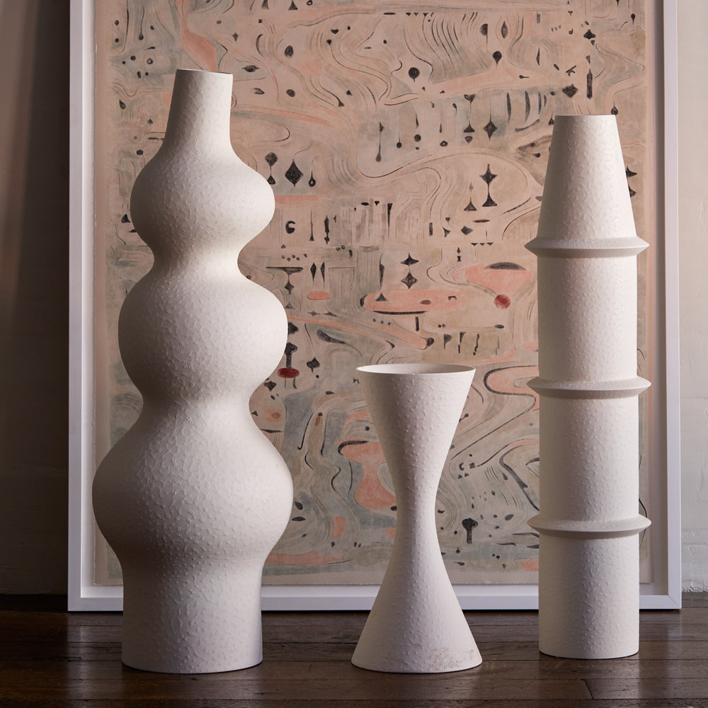 Lemieux et Cie Deoune Vase in matte white, sculptural ceramic vase with organic bulbous form, displayed with other vases against abstract art on wood flooring.