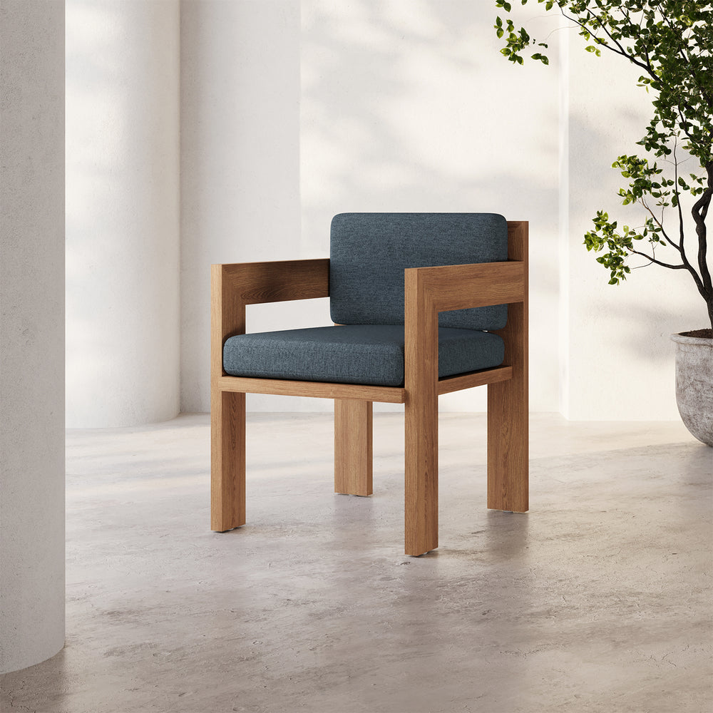 Lemieux et Cie S2 Teak Dining Arm Chair with deep blue-gray cushions, angular outdoor dining chair on stone floor in white courtyard with greenery.