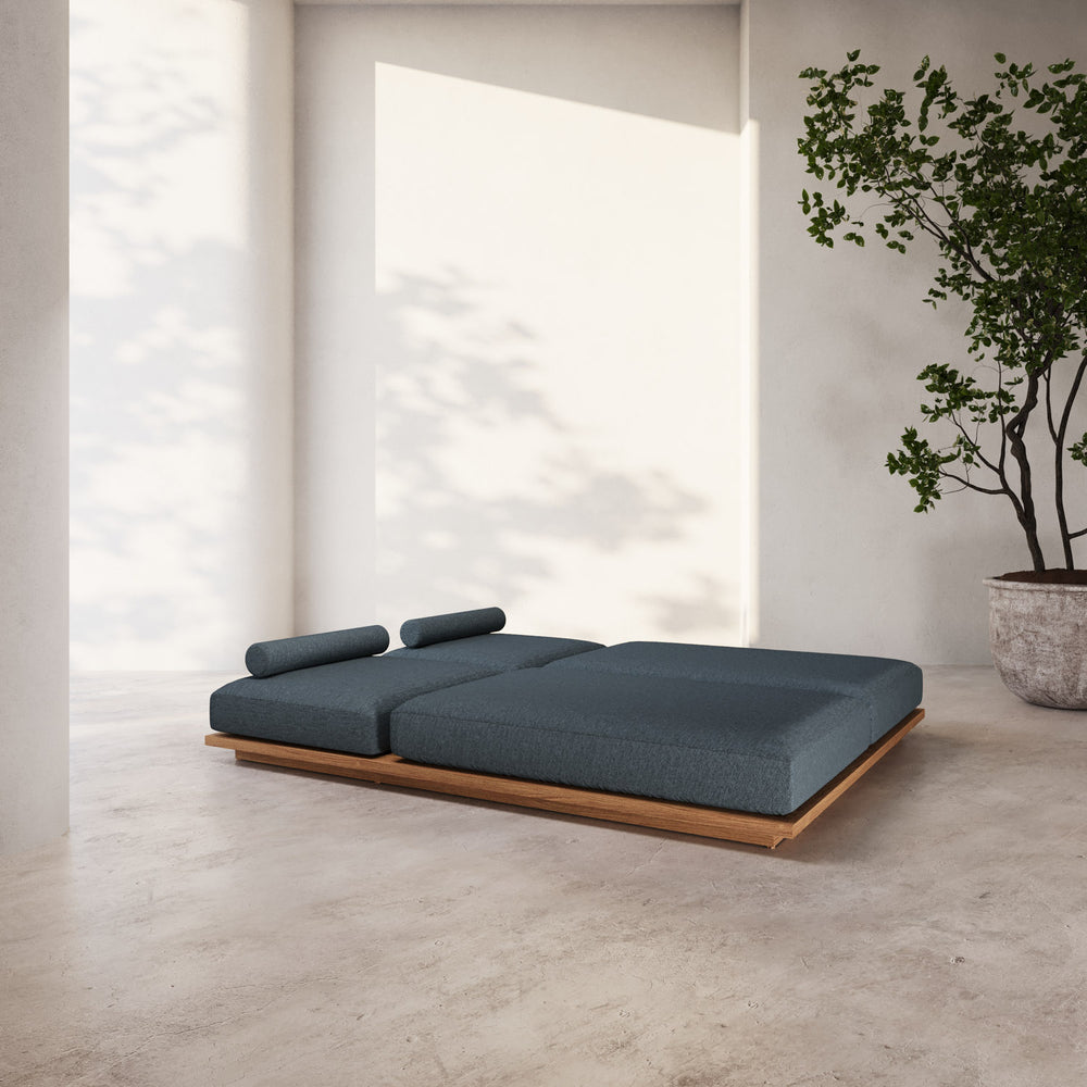 Lemieux et Cie S2 Teak Double Chaise Lounge in deep blue-gray cushioning on solid teak base, shown on concrete floor in light-filled room with white walls.