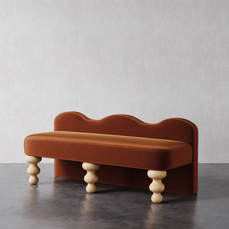 Lemieux et Cie Celine Bench in warm terracotta upholstery with sculptural undulating backrest and turned cream legs, shown on dark concrete floor against white plaster wall.