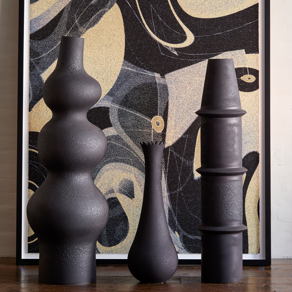 Lemieux et Cie black vases in varied organic sculptural forms, grouped on wood flooring against framed abstract artwork in charcoal and sand tones.