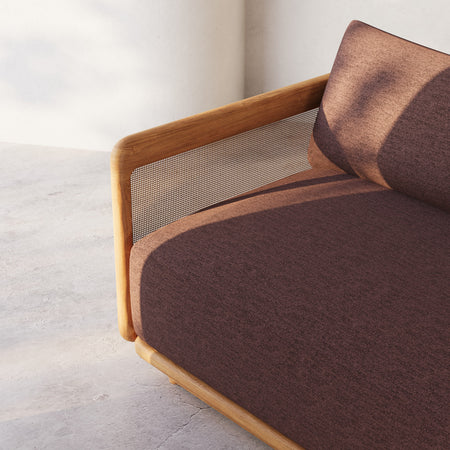 Lemieux et Cie Dock Teak Chaise Lounge with caned back panel and warm brown cushions, low-profile outdoor seating on concrete floor against white walls.