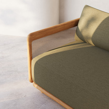 Lemieux et Cie Dock Teak Lounge Chair in olive-green upholstery with woven cane panels and rounded teak frame, shown on concrete floor against white walls.