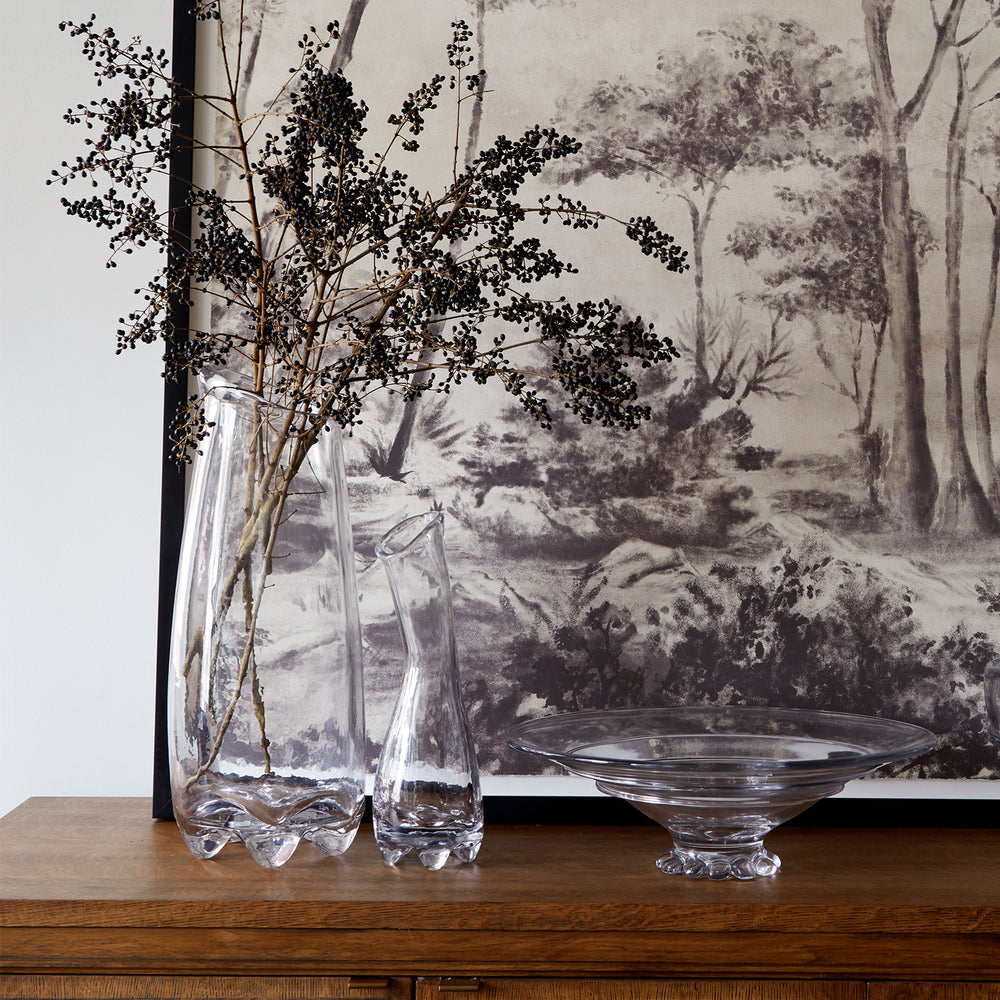 Lemieux et Cie Fleur de Verre glass vase with organic curved form, styled with dark branches on wood surface against hand-painted landscape mural.