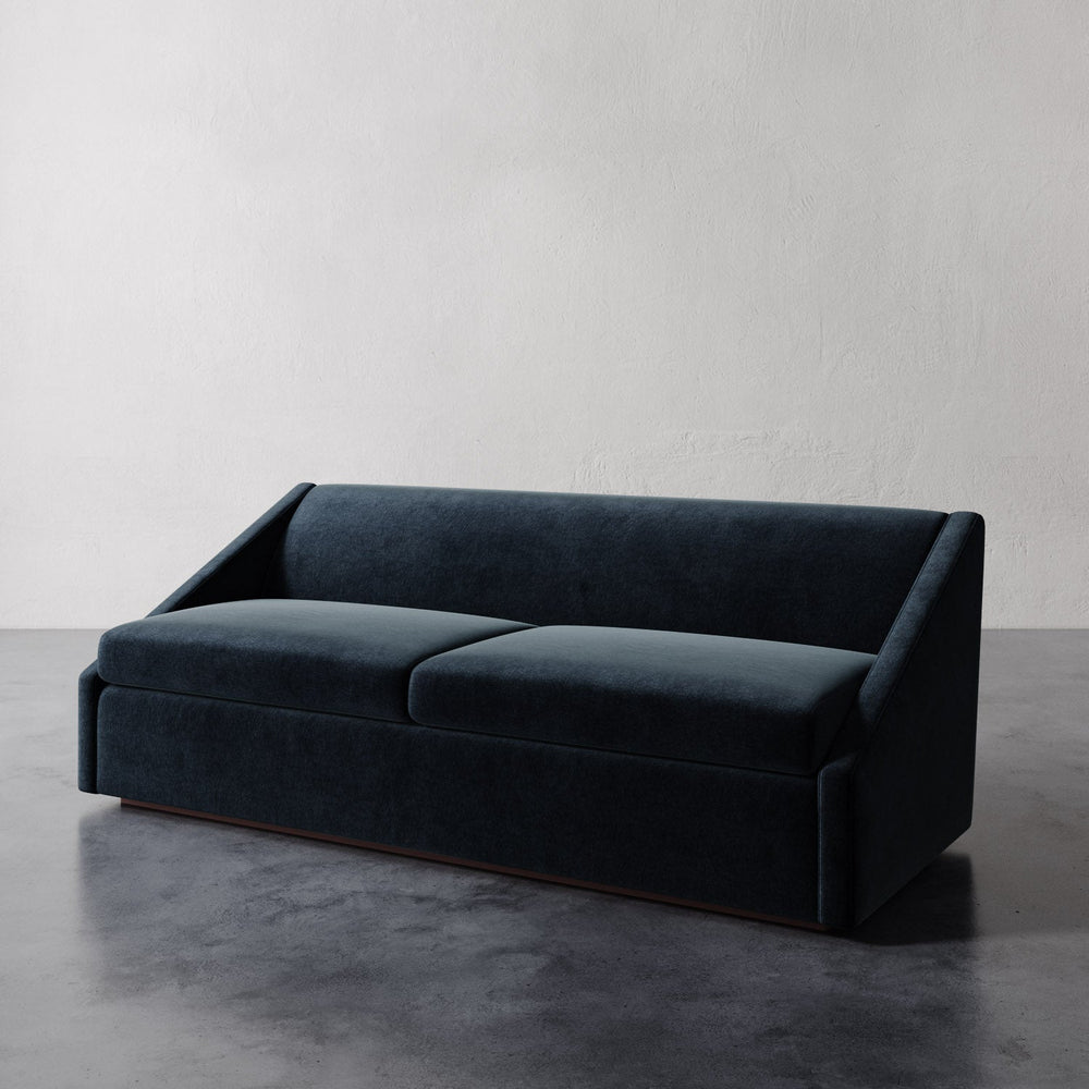 Lemieux et Cie Fontaine Sofa in dark upholstery, architectural low-profile sofa with angled arms, shown on polished concrete floor against white plaster walls.