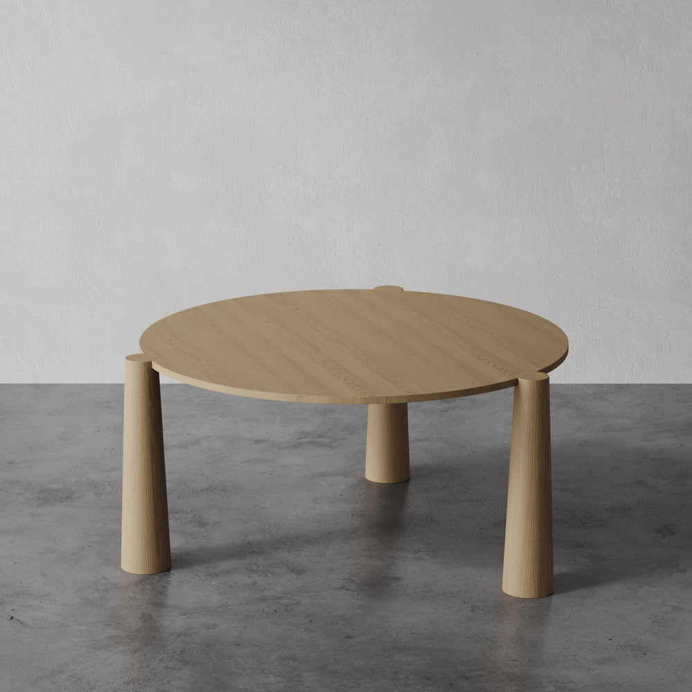 Lemieux et Cie Girard Dining Table in oak with flared cylindrical legs, low-profile coffee table on concrete flooring against neutral walls.
