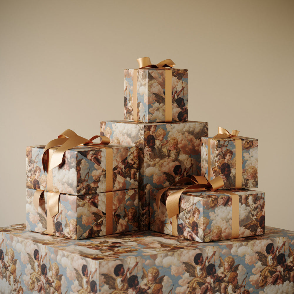 Lemieux et Cie Grecian Cherub Gift Wrapping Paper with celestial figures and clouds in soft blue and rose tones, stacked boxes tied with gold ribbon against neutral backdrop.