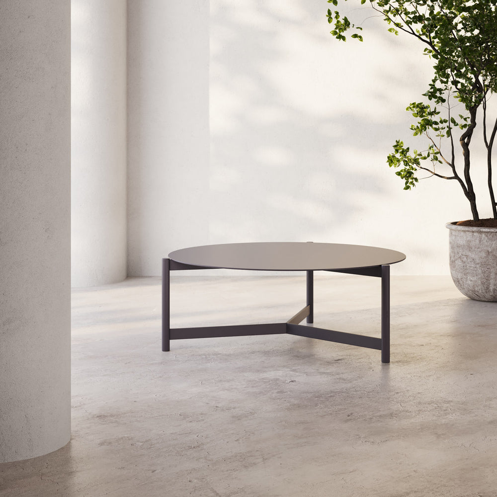 Lemieux et Cie Hudson 35" Round Coffee Table, low-profile round form with cross-base, shown on concrete flooring in soft daylight beside potted tree.