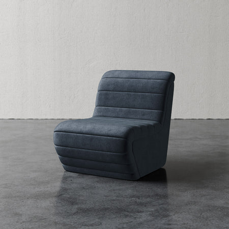 Lemieux et Cie Brunoy Chair in charcoal, channel-tufted lounge chair with curved low-back silhouette, shown on polished concrete floor against textured plaster wall.