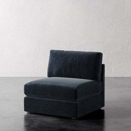Lemieux et Cie Chanaux Armless Chair in deep charcoal upholstery, slipper chair shown on dark concrete floor against textured white plaster wall.