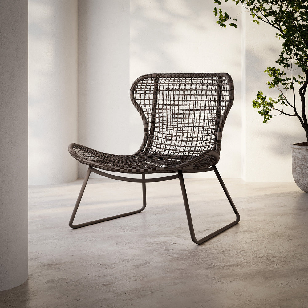 Lemieux et Cie Jagger Woven Lounge Chair with open-weave seat and back on sled base, outdoor lounge chair on stone floor near white columns and potted tree.