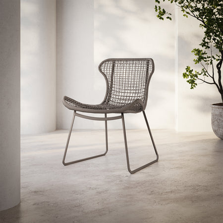 Lemieux et Cie Jagger Woven Dining Chair with open-weave seat and back on sled base, shown on concrete floor against white columns in dappled daylight.