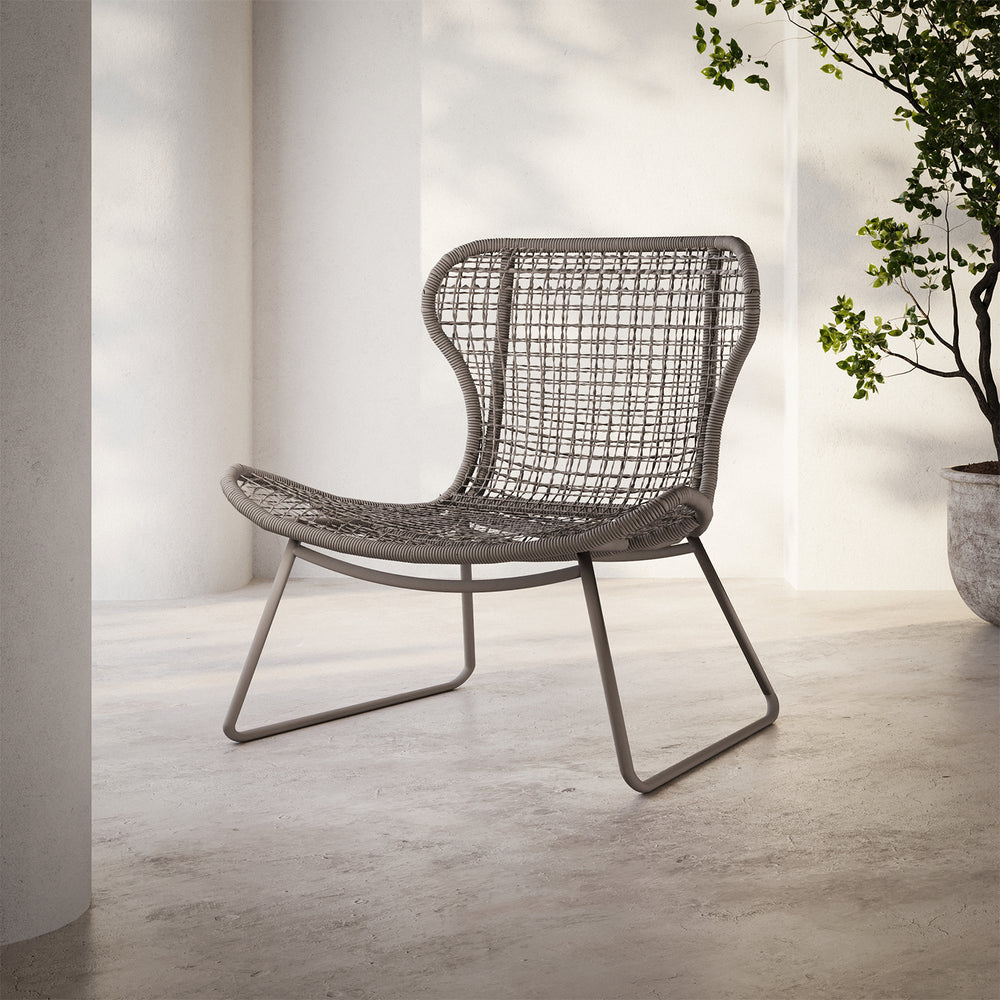 Lemieux et Cie Jagger Woven Lounge Chair with open-weave seat and back on sled base, shown on concrete floor near white columns and greenery.