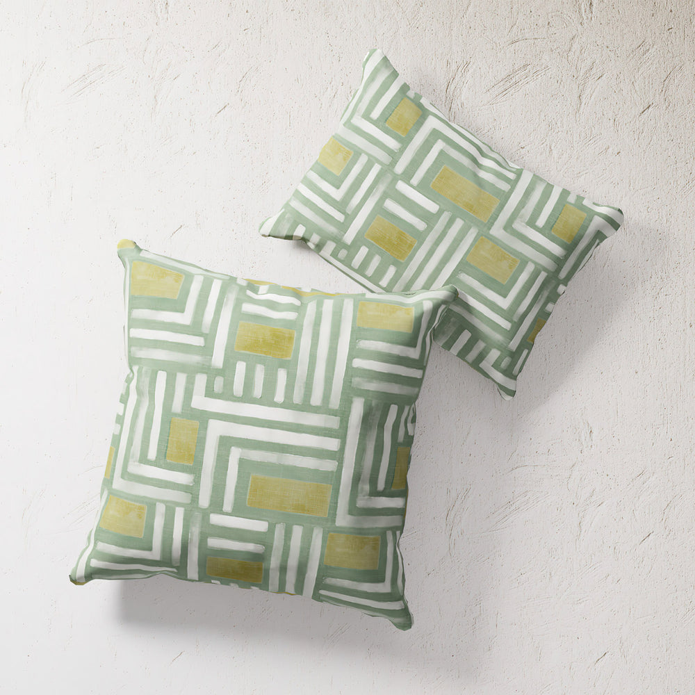 Lemieux et Cie Painterly Labyrinth Outdoor Pillow in green and yellow geometric pattern, double-sided cushion shown on textured plaster surface