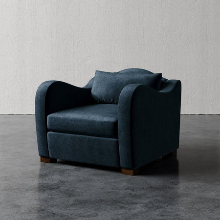 Lemieux et Cie Elodie Chair in deep blue upholstery, low-profile lounge chair with rounded arms, shown on polished concrete floor against textured white plaster wall.