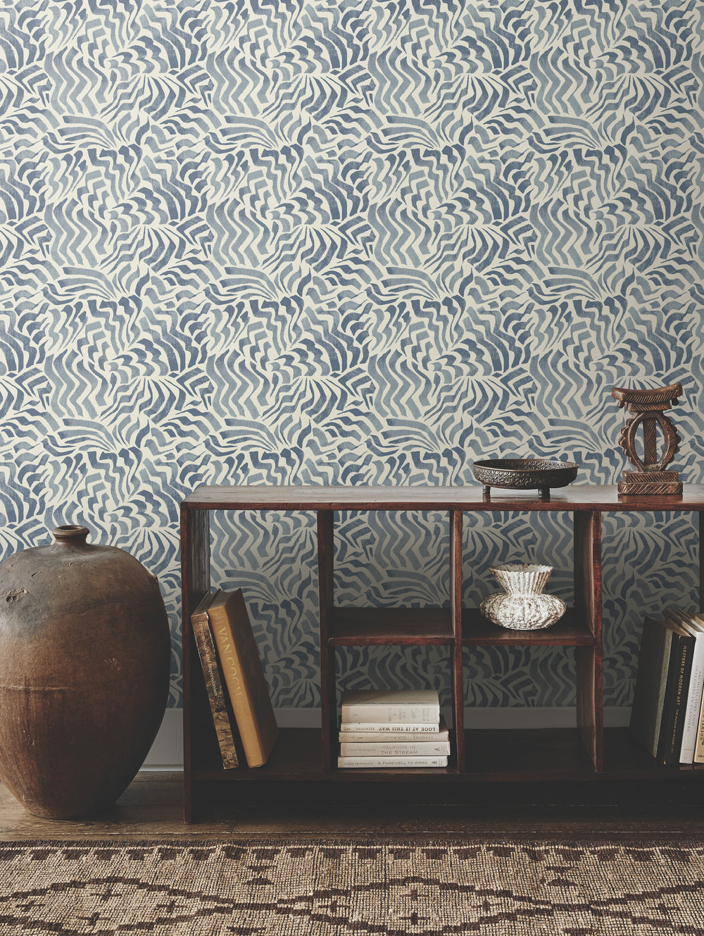 Lemieux et Cie Zora Waves wallpaper in indigo blue on white, featuring organic wave motifs, styled with wood shelving and ceramic vessels on woven rug.