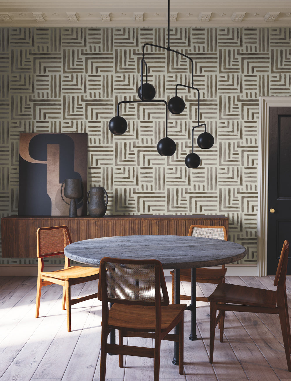 Lemieux et Cie Painterly Labyrinth wallpaper in taupe and brown geometric pattern, dining room with round stone table, caned chairs, matte spherical chandelier, pale wood flooring.