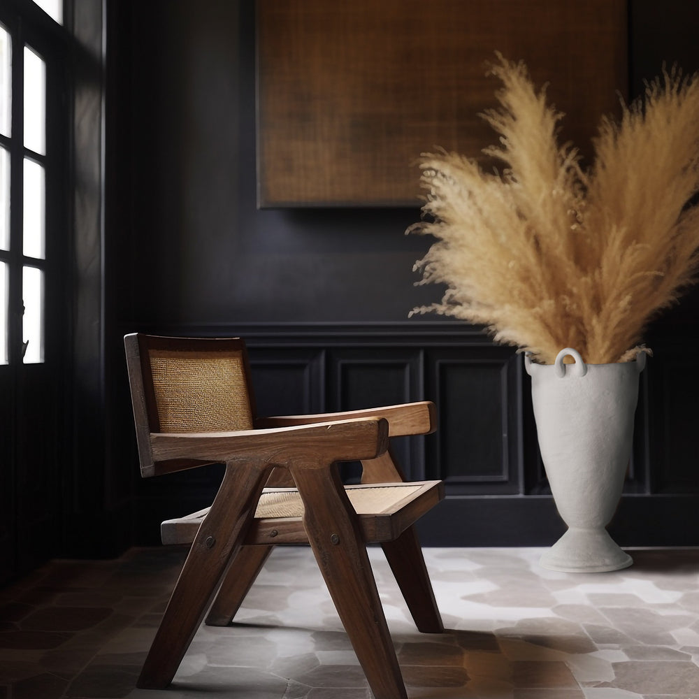 Lemieux et Cie Louis Vase in white, sculptural ceramic vessel with organic form and handles, styled with pampas grass beside a caned lounge chair in dark-paneled room with stone floor.