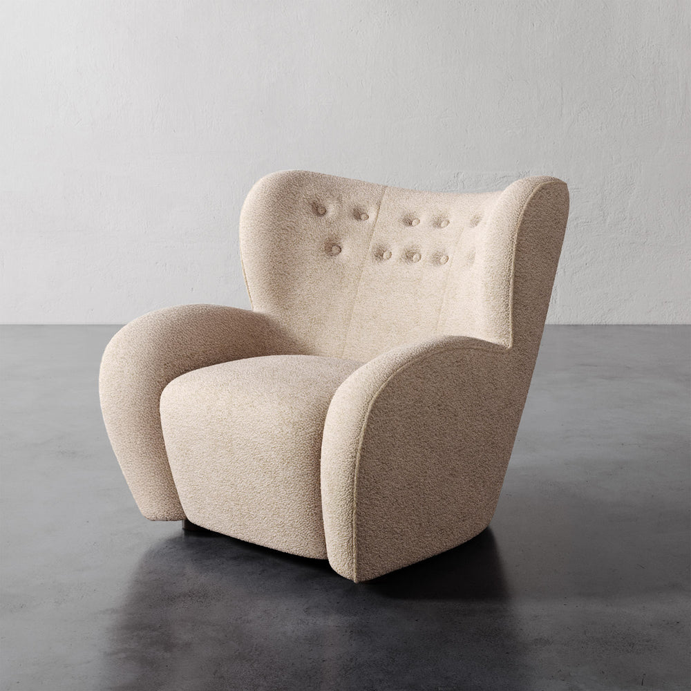 Lemieux et Cie Mondaine Lounge Chair in neutral bouclé, sculptural wingback chair with button-tufted back, set on dark concrete floor against white textured walls.