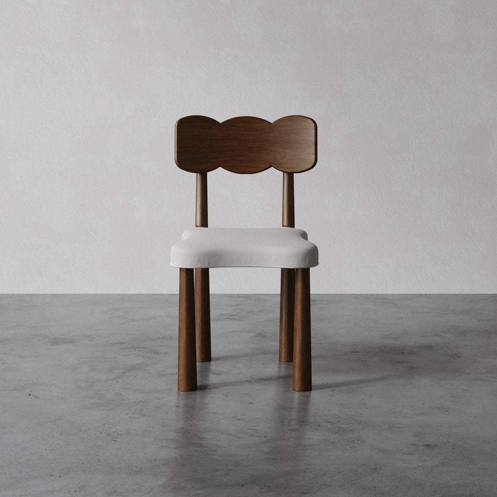 Lemieux et Cie dining chair in oak with scalloped back and ivory upholstered seat, shown on concrete floor against neutral plaster walls.
