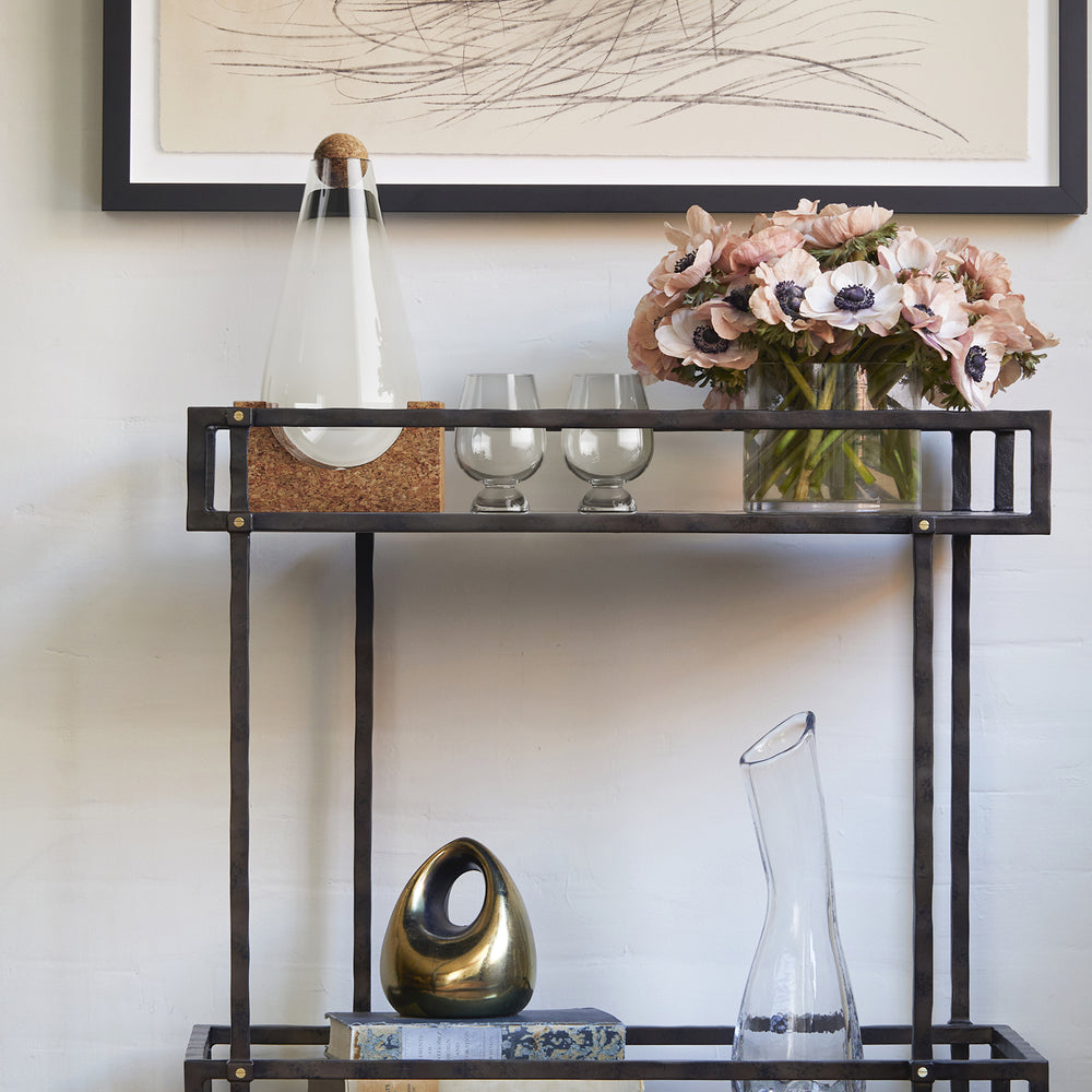 Lemieux et Cie glass decanter with geometric cork base and spherical stopper, styled on dark open-frame console against white walls with abstract artwork.