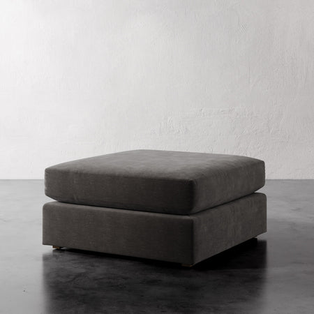 Lemieux et Cie Chanaux Ottoman, low-profile rectangular cushioned ottoman in charcoal, shown on dark concrete floor against textured white plaster walls.