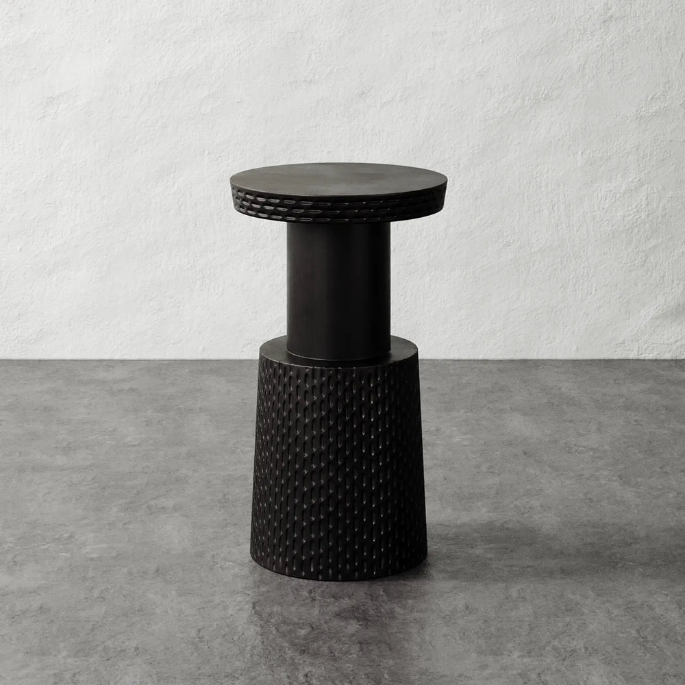 Lemieux et Cie Morel Pedestal Side Table in dark textured resin with hand-worked grooves, sculptural side table on concrete floor against white plaster wall.