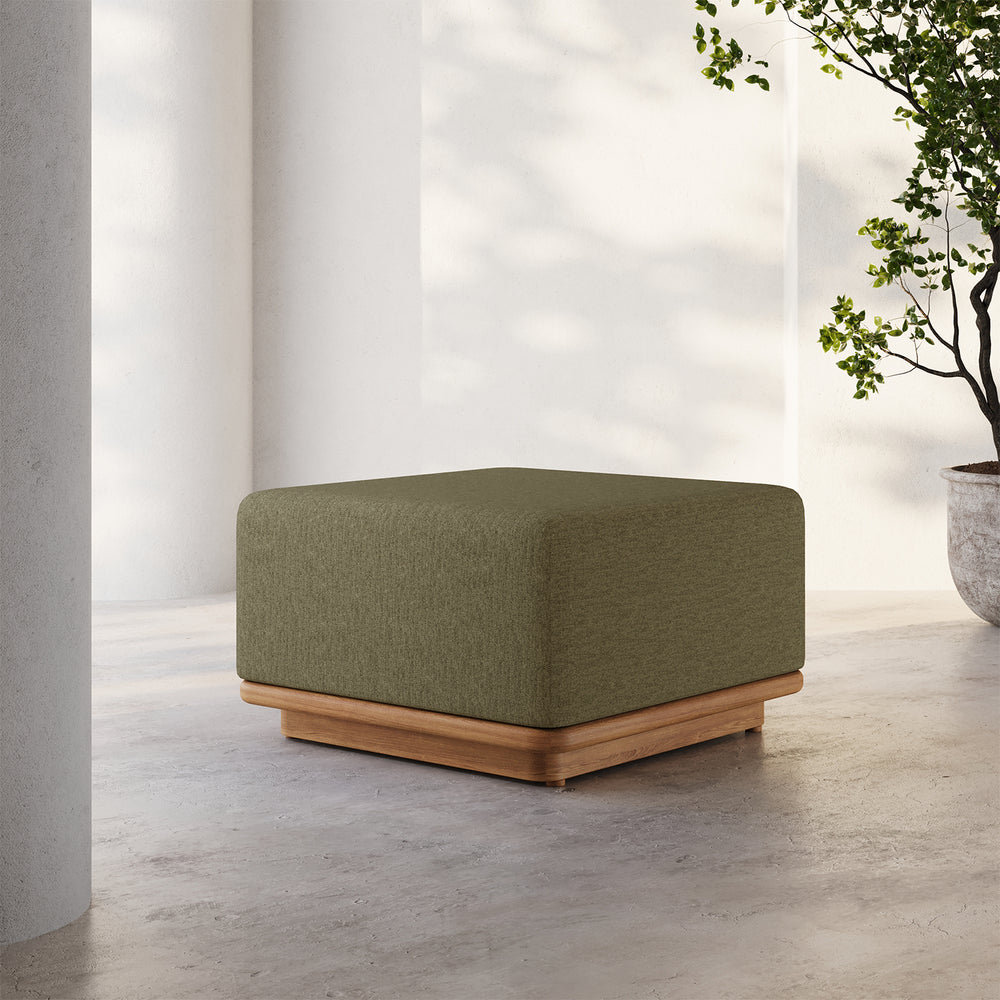 Lemieux et Cie Dock Teak Ottoman in olive green upholstery on teak base, shown on concrete floor in bright courtyard with white walls.