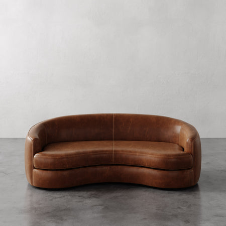 Lemieux et Cie Rulmann Sofa in warm cognac leather, curved organic form with rounded arms, shown on concrete floor against neutral plaster walls.