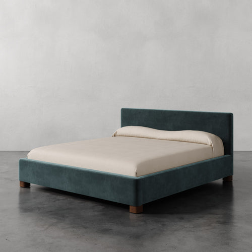 Lemieux et Cie Gérard Bed in teal upholstery with ivory linens, low-profile platform bed on dark concrete floor in neutral plaster-walled room.