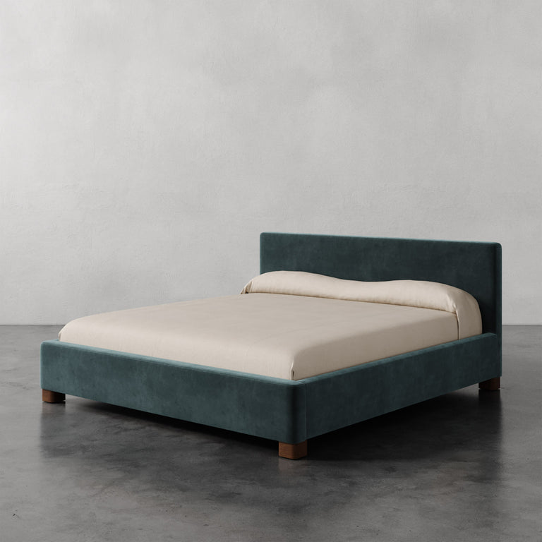 Lemieux et Cie Gérard Bed in teal upholstery with ivory linens, low-profile platform bed on dark concrete floor in neutral plaster-walled room.
