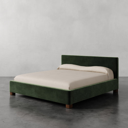 Lemieux et Cie Gérard Bed in deep green upholstery, low-profile platform bed with wrapped headboard and block feet, on concrete floor against neutral plaster walls.