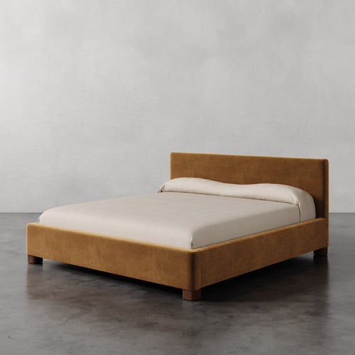 Lemieux et Cie Gérard Bed in warm caramel upholstery, low-profile platform bed with wrapped headboard, shown on polished concrete floor against neutral plaster walls.