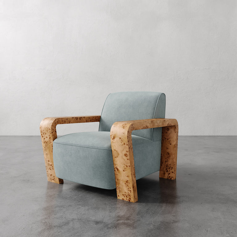 Lemieux et Cie Dumas Chair in pale blue upholstery with sculpted blonde burl arms, lounge chair on polished concrete floor against textured plaster wall.