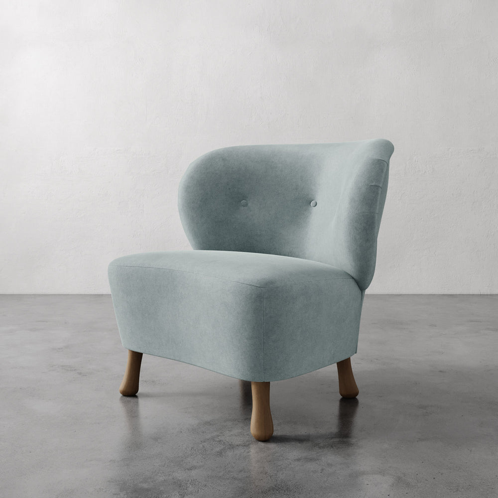 Lemieux et Cie Tourenne Chair in soft sage upholstery with rounded back and button tufting, sculptural slipper chair on turned legs, shown on concrete floor against neutral wall.