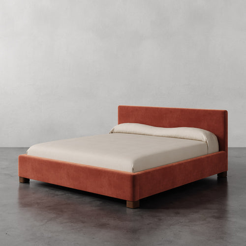 Lemieux et Cie Gérard Bed in terracotta upholstery, low-profile platform bed with rounded block feet, shown on polished concrete floor against neutral plaster walls.
