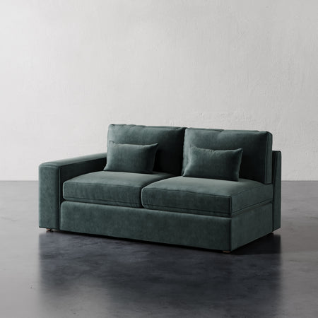 Lemieux et Cie Lacroix Left or Right Arm Loveseat in deep green velvet, modular sofa with rectilinear form and bolster pillows, on dark concrete floor.