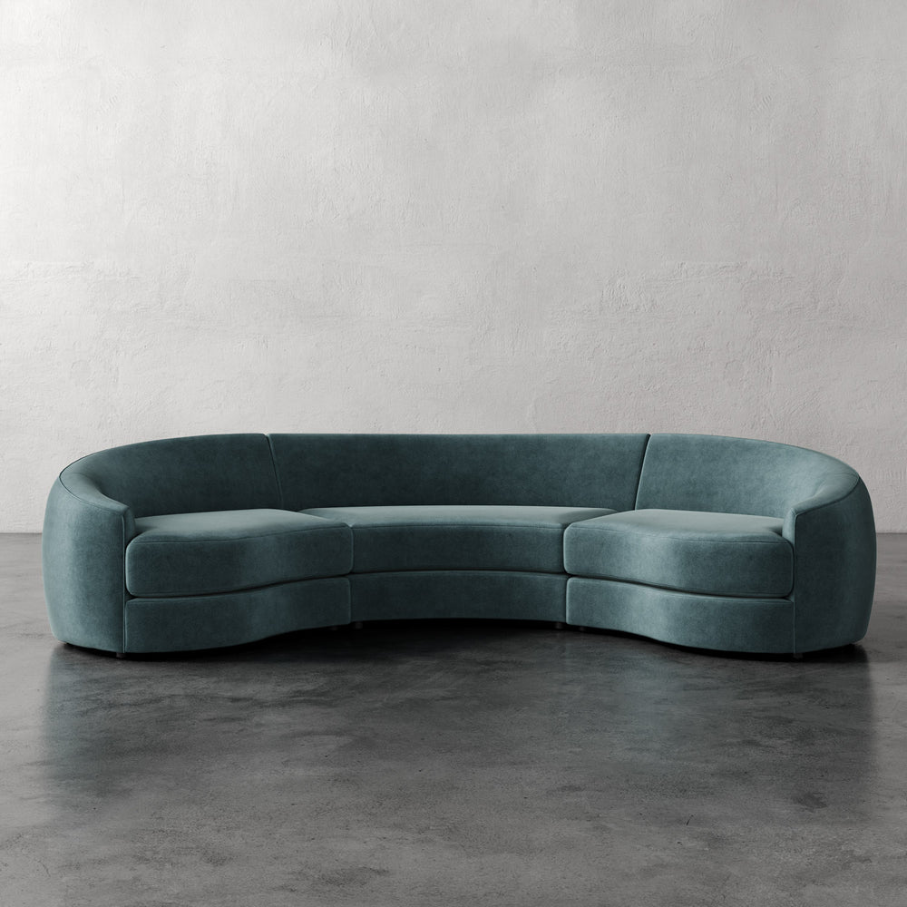 Lemieux et Cie Rulmann 3-Piece Sectional in teal, curved sculptural sofa with rounded arms, shown on dark concrete floor against textured plaster walls.