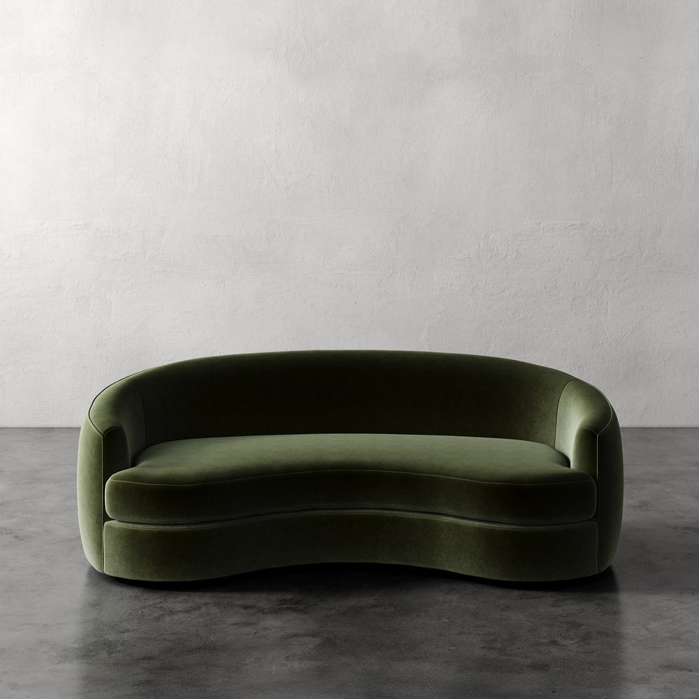 Lemieux et Cie Rulmann Sofa in deep olive green, curved sculptural sofa with rounded arms, set against textured plaster walls and dark concrete floor.