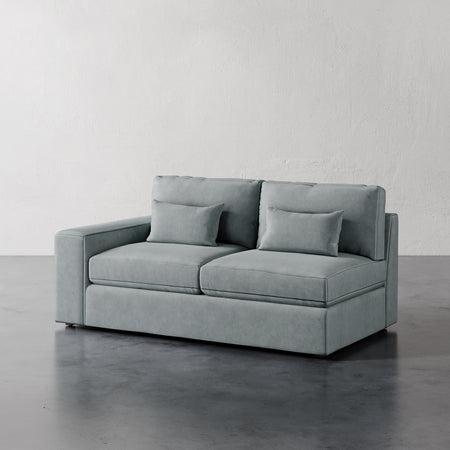 Lemieux et Cie Lacroix Left or Right Arm Loveseat in soft gray-blue, modular sofa with bolster pillows on dark concrete floor against white walls.