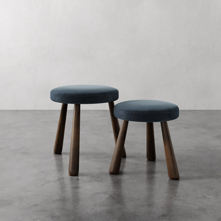 Lemieux et Cie Madeleine Stool with dark blue upholstered seat and tapered legs, pair of stools shown on polished concrete floor against neutral plaster walls.