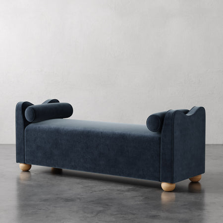 Lemieux et Cie Danielle Upholstered Bench in charcoal blue with cylindrical arm bolsters and turned legs, shown on polished concrete floor against neutral plaster walls.