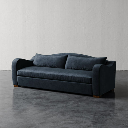 Lemieux et Cie Elodie Sofa in deep charcoal, bench seat sofa with rounded arms and single cushion, shown on polished concrete floor against textured plaster walls.