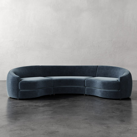 Lemieux et Cie Rulmann 3-Piece Sectional in deep blue-gray, curved sculptural sofa with rounded arms, shown on polished concrete floor against textured plaster walls.