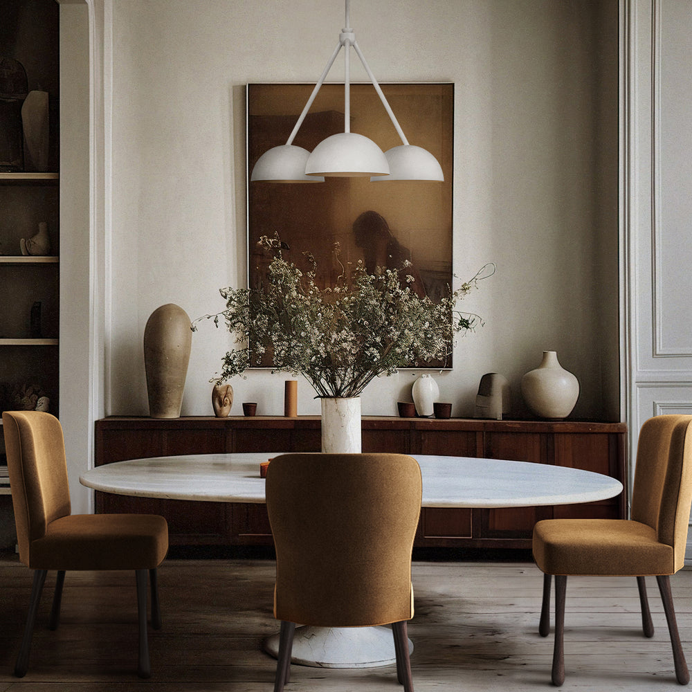 Lemieux et Cie Beaunay Medium Chandelier, a luxury lighting fixture in steel and cast plaster with three bowl-shaped shades, glowing softly above a circular dining table with Rozette Dining Chairs in a sunlit dining room.