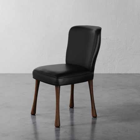 Lemieux et Cie Rozette Dining Chair in black leather with tapered legs, shown on polished concrete floor against textured white walls.