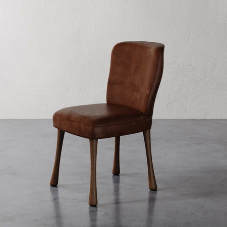 Lemieux et Cie Rozette Leather Dining Chair in warm brown leather with tapered legs, shown on polished concrete floor against textured white wall.