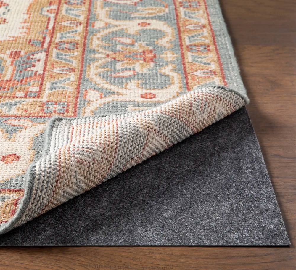 Lemieux et Cie Lemieux et Rie Standard Felted Rug Pad in charcoal felt, shown layered beneath a patterned rug with corner folded to reveal cushioned backing on wood floor.