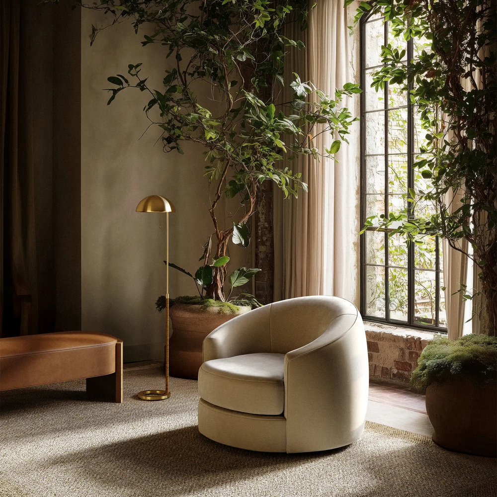Lemieux et Cie Benoit Leather Bench, rounded low-profile bench in warm caramel tone, shown beside swivel chair and brass floor lamp in sitting room with plaster walls and tall windows.
